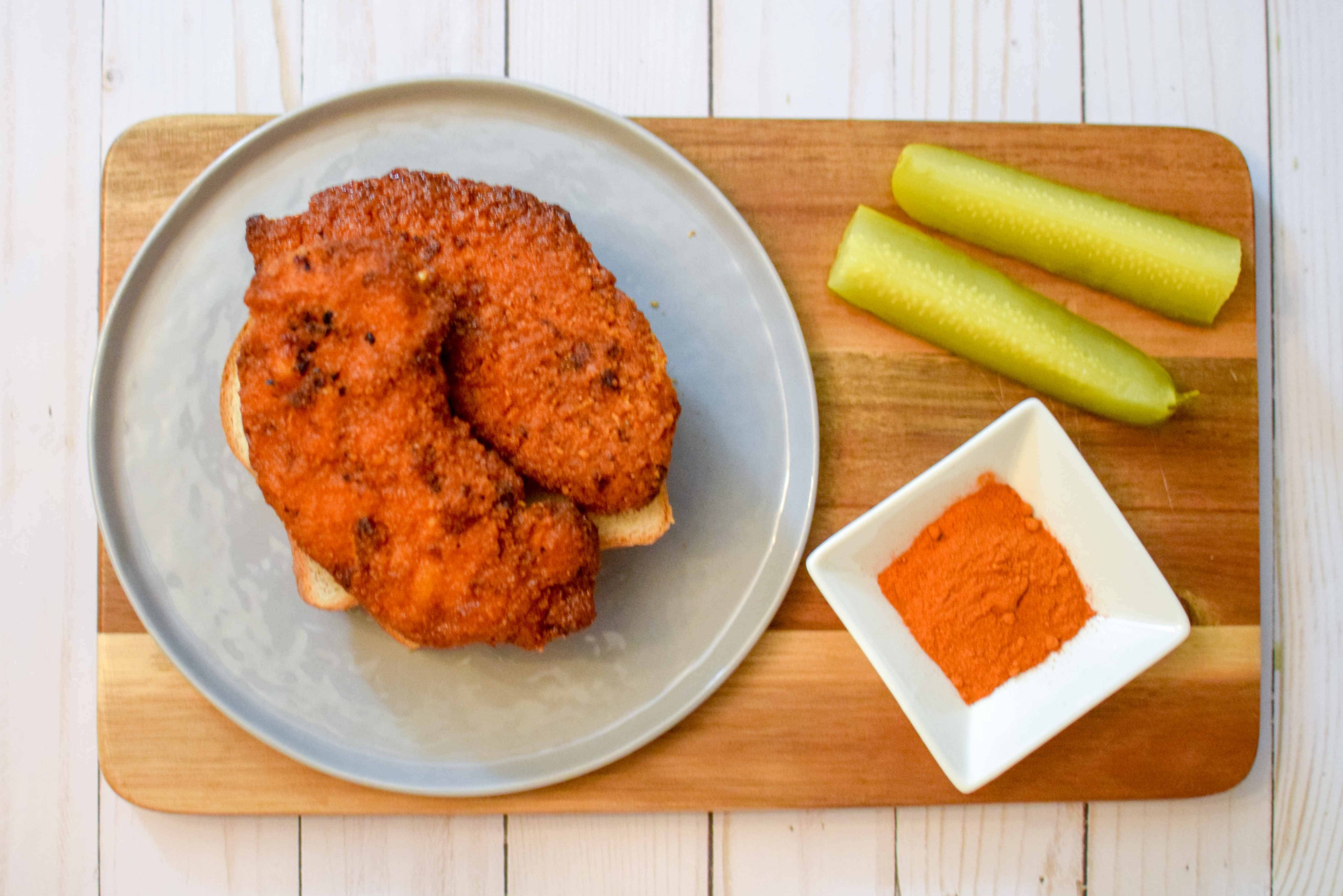 Two pieces of finished crispy chicken on a grey plate with pickles and seasoning blend. 