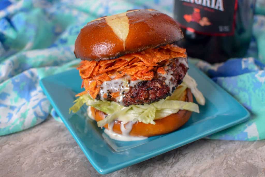 Blazin Queso Burger with a pretzel bun topped with spicy doritos and queso cheese sauce served on a square blue plate.