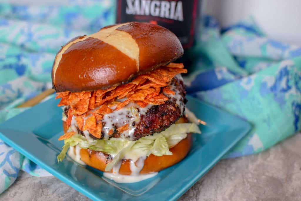 Burger with a pretzel bun topped with spicy Doritos and queso cheese sauce served on a square blue plate with a bottle of Kirkland Sangria behind it on a decorative blue towel.