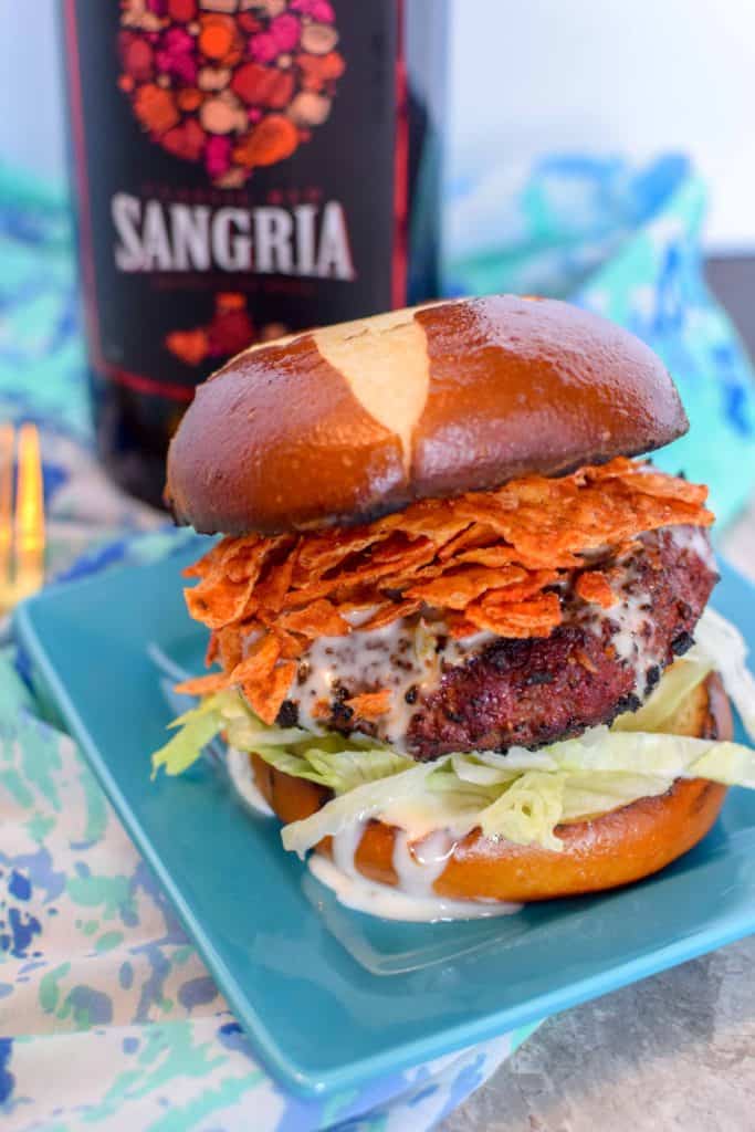 Burger with a pretzel bun topped with spicy Doritos and queso cheese sauce served on a square blue plate with a bottle of Kirkland Sangria behind it.