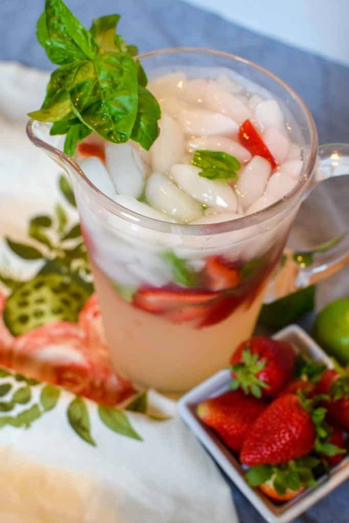A pitcher full of Strawberry Basil Margaritas with fresh cut strawberries and basil inside, next to a dish of fresh whole strawberries.