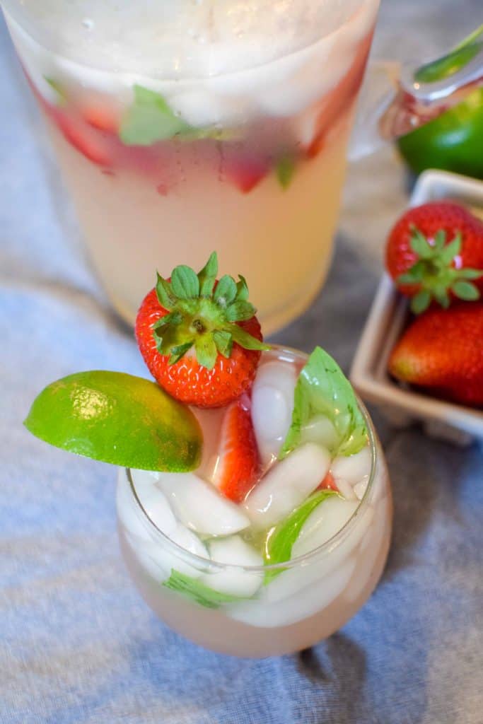 Easy Strawberry Basil Margarita in a wine glass with ice and topped with a lime wedge and fresh strawberry, with a pitcher and fresh strawberries in the background.