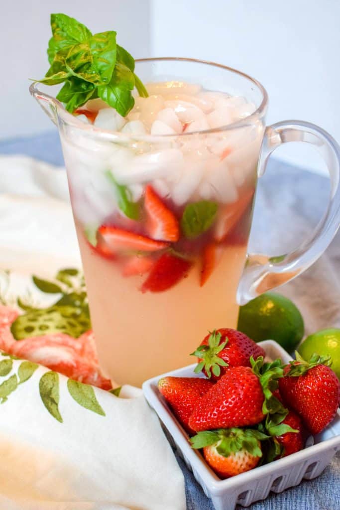 A pitcher full of Strawberry Basil Margaritas with fresh cut strawberries and basil inside, next to a dish of fresh whole strawberries.