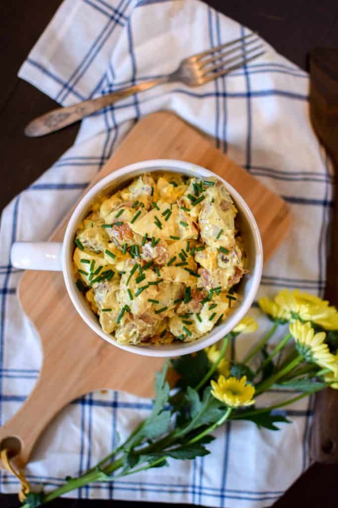 Red Skinned Potato Salad in a white soup cup with yellow flowers and a fork next to the serving platter.