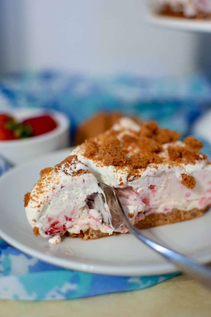 Slice of Strawberry Ice Cream Cake on a white plate having a metal fork slice a small piece off.