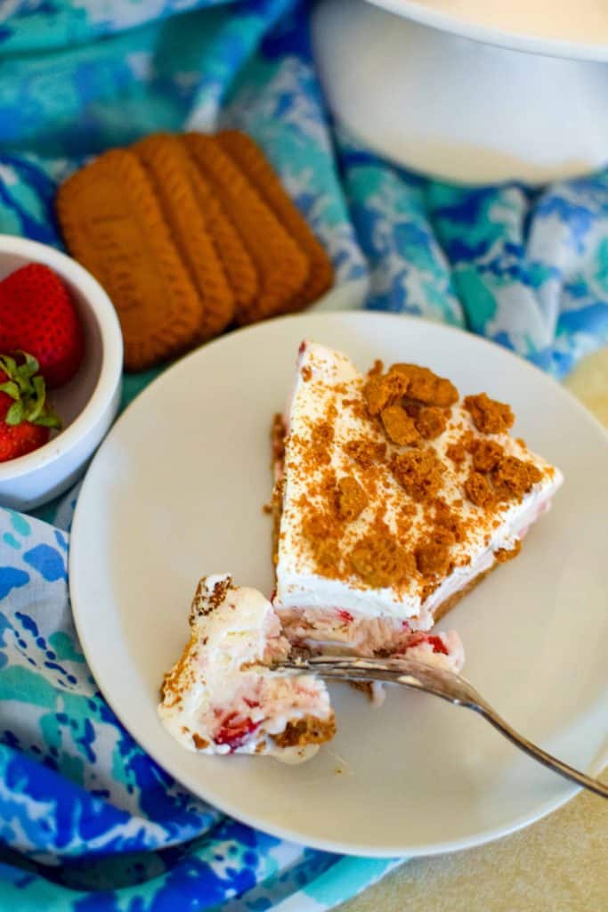 Fork cutting through a slice of Strawberry Ice Cream Cake on a white plate with biscoff cookies next to it.