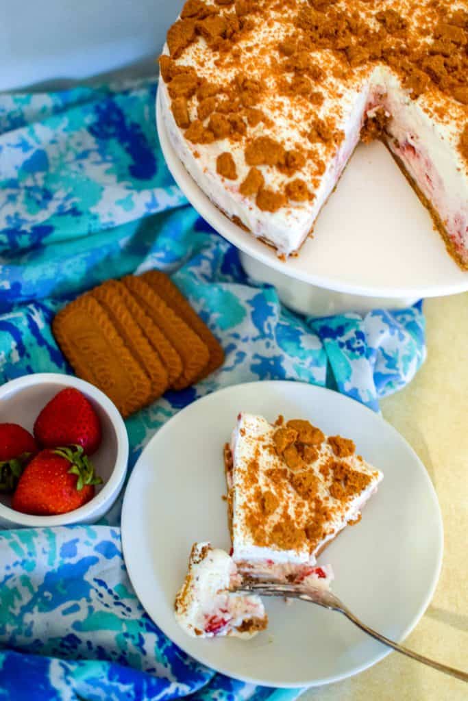 Whole Strawberry Ice Cream Cake with a Biscoff Cookie Crust shown next to a slice of the cake on a small white plate with a fork cutting into it.
