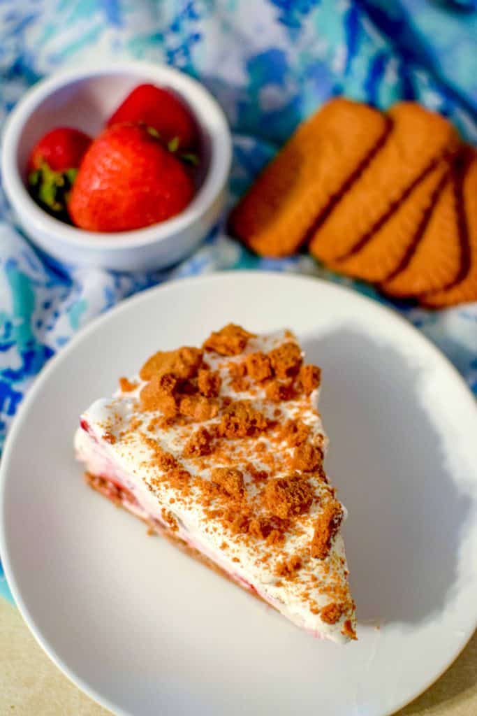 Slice of Strawberry Ice Cream Cake on a white plate with fresh strawberries and Biscoff cookies on a decorative blue towel in the background.