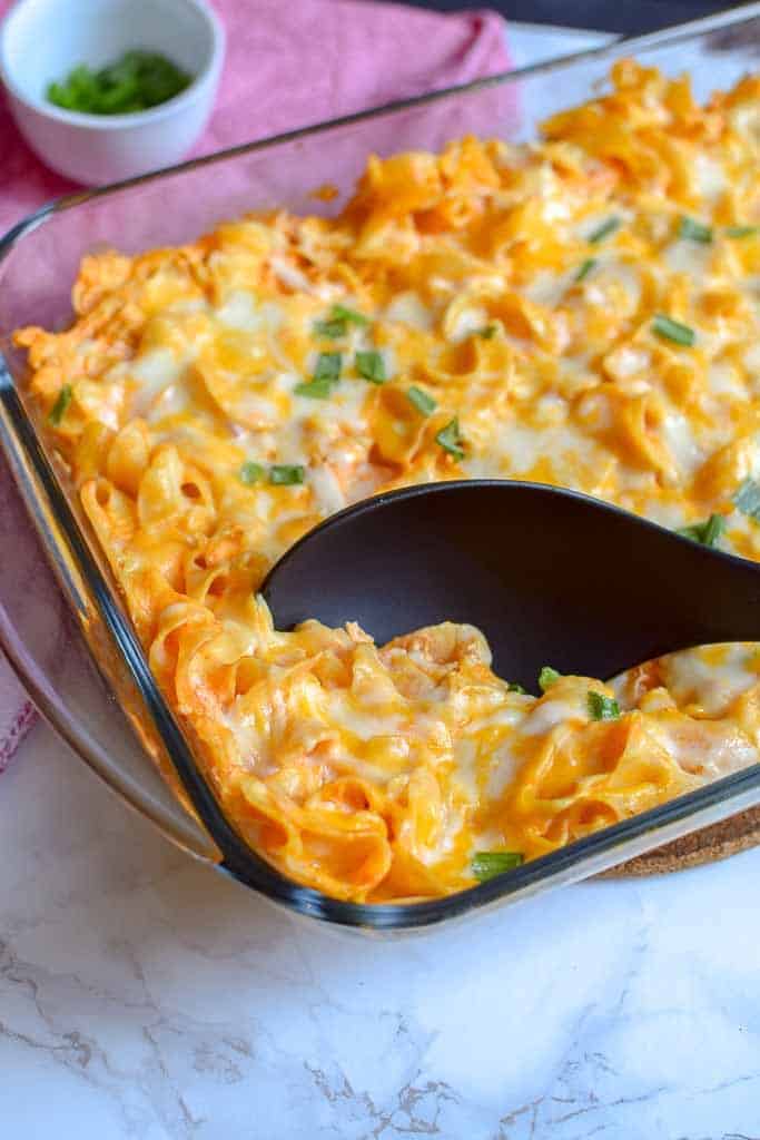 Buffalo Chicken Casserole in a baking dish being scooped out by a large spoon with a cup of chopped green onions in the background.