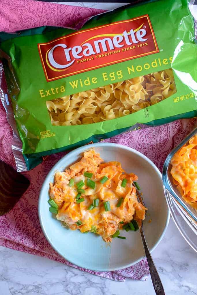 Homemade buffalo chicken casserole shown in a white bowl with a metal fork alongside a green bag of Creamette extra wide egg noodles.