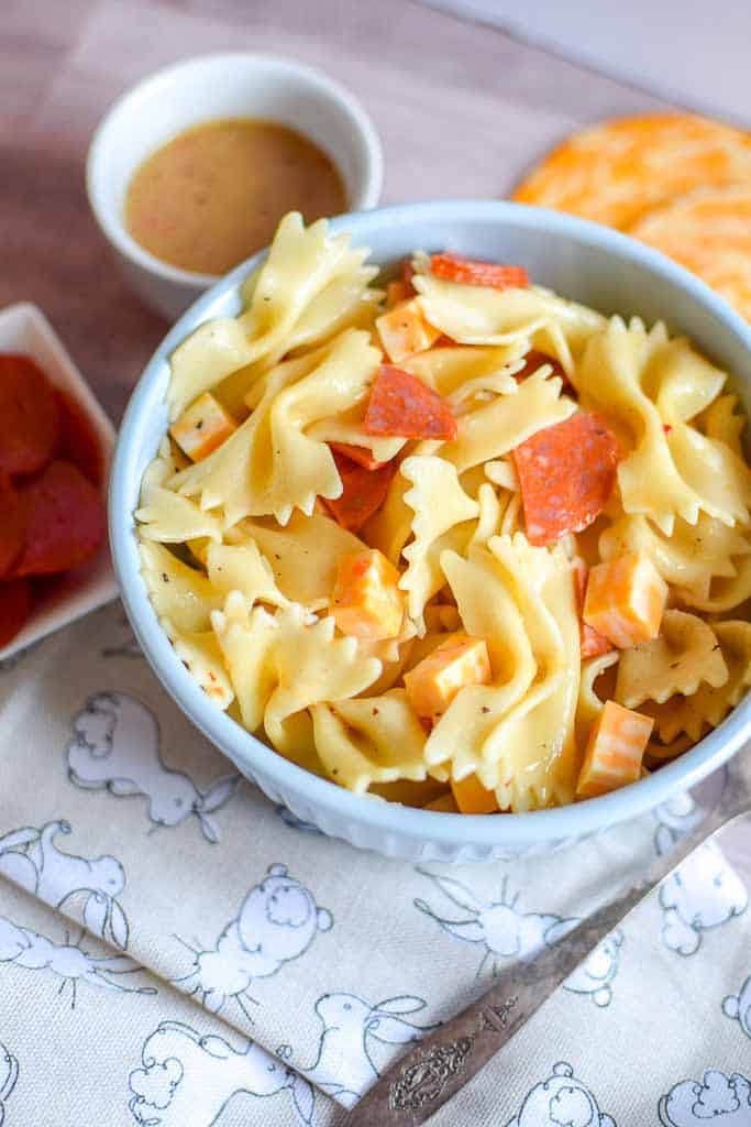 4 ingredient Pasta Salad shown in a bowl with a side dish of pepperoni and a cup of dressing.