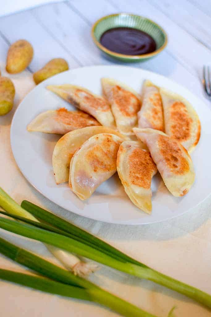 A white plate filled with corned beef dumplings with a small dish of bbq sauce in the background and whole green onions in the foregorund.