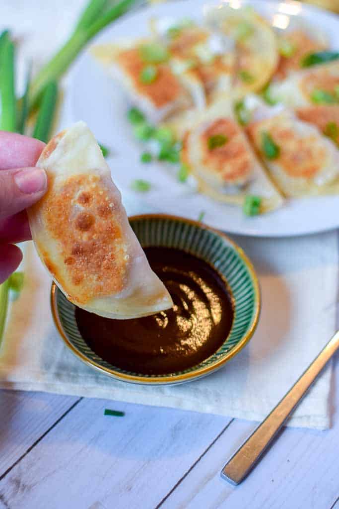 Dumpling being dipped into a dish of barbecue sauce with a plate full of dumplings in the background.