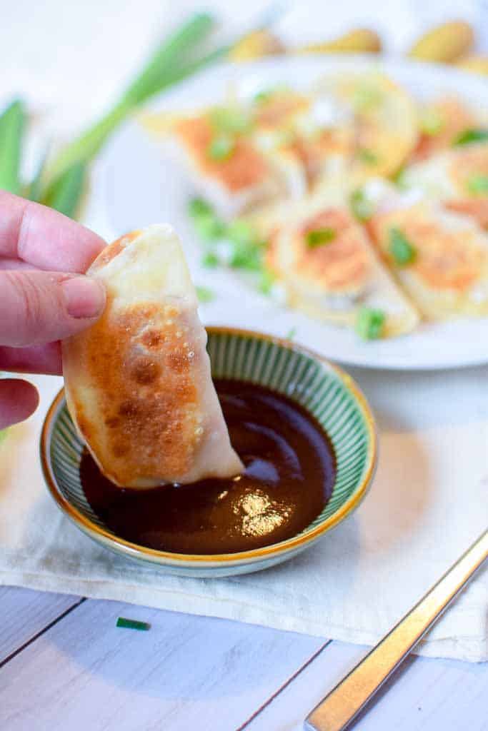 A hand holding a corned beef dumpling and dipping it into a dish of guinness stout bbq sauce with a gold fork next to the dish.