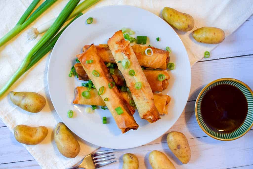A plate full of stacked corned beef spring rolls topped with chopped green onions with a fork, cup of bbq dipping sauce, and whole green onions next to the plate.