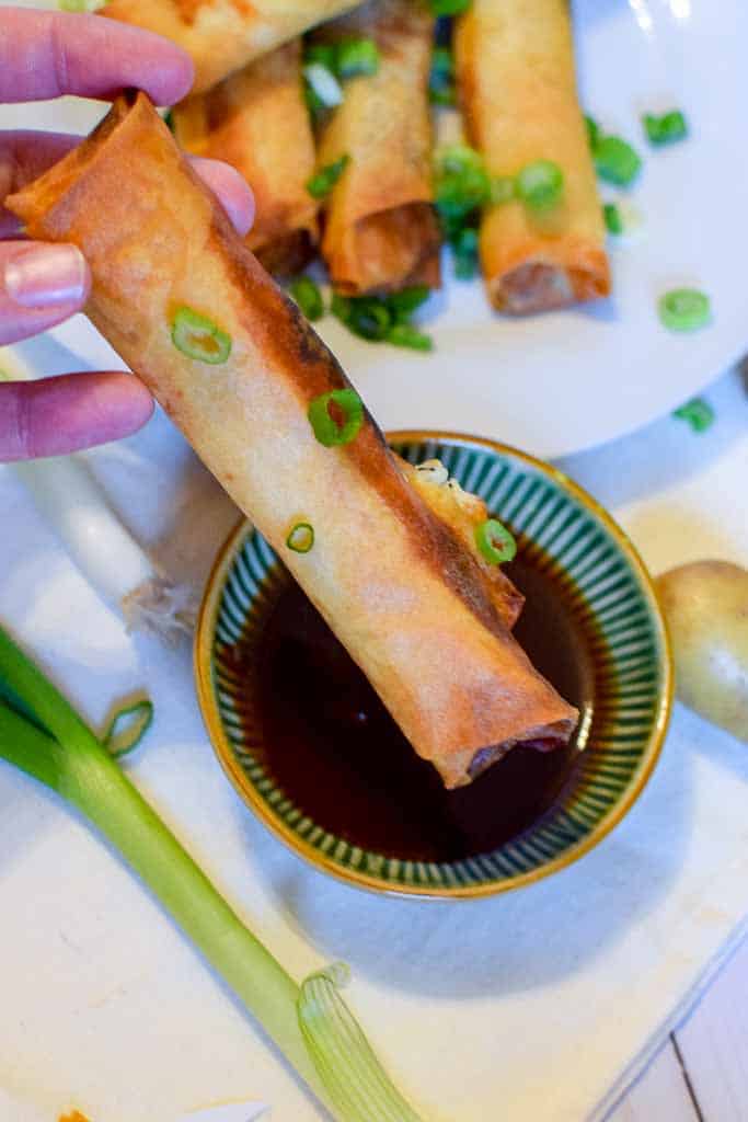 A corned beef spring roll topped with green onions and being dipped into homemade bbq sauce.