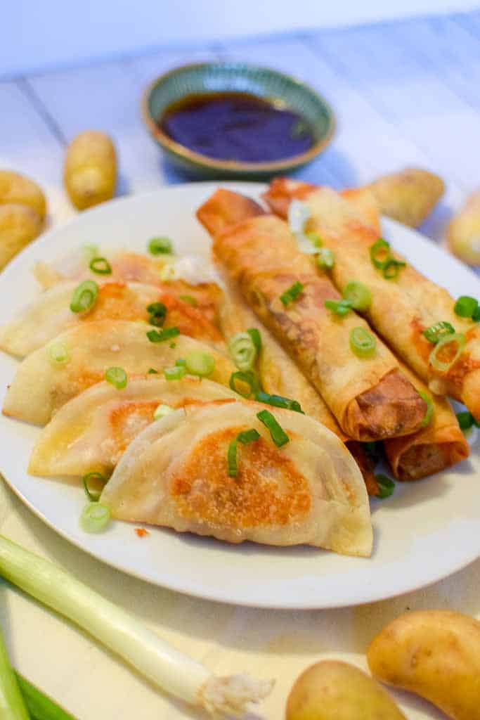A white plate filled with corned beef dumplings and spring rolls, topped with green onions, in front of a dish of bbq sauce.