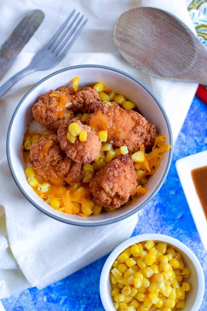 KFC Famous Bowl copycat shown in a white bowl with a blue rim, sitting on a white napkin with a fork and wooden spoon, next to a side bowl of golden sweet corn.