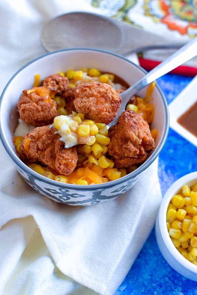Mashed potatoes, sweet corn, and crispy chicken with gravy on a fork, over top of a kfc famous bowl on a white napkin and blue table top, with a wooden spoon in the background.