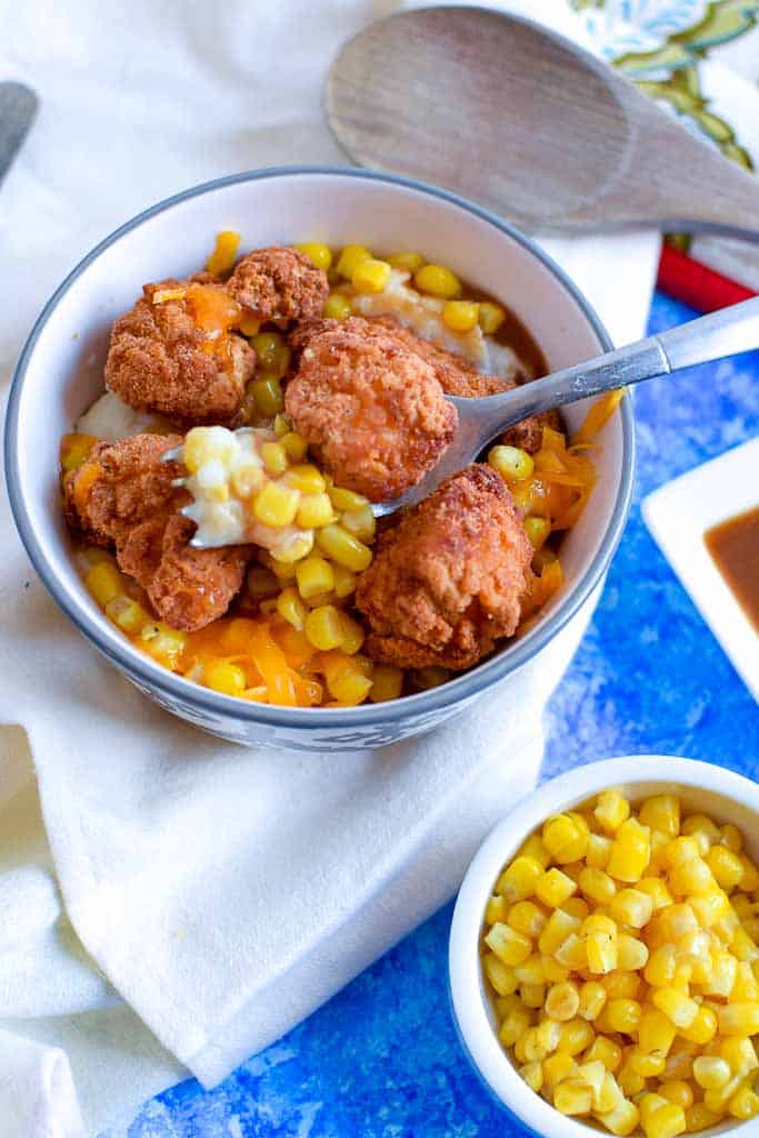 Mashed potatoes, sweet corn, and crispy chicken with gravy on a fork over top of a kfc famous bowl on a white napkin and blue table top, with a side dish of sweet corn.