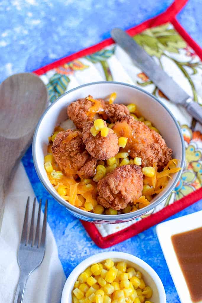 Closeup of copycat KFC Famous bowl of mashed potatoes topped with sweet corn, gravy, and cheddar cheese, on top of a pot holder, with a side dish full of sweet corn and a fork.