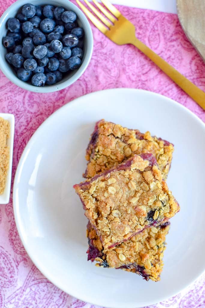 Incredible Berry, Rhubarb, And Oat Breakfast Bar squares on a white plate with a small bowl of fresh blueberries and a gold fork.