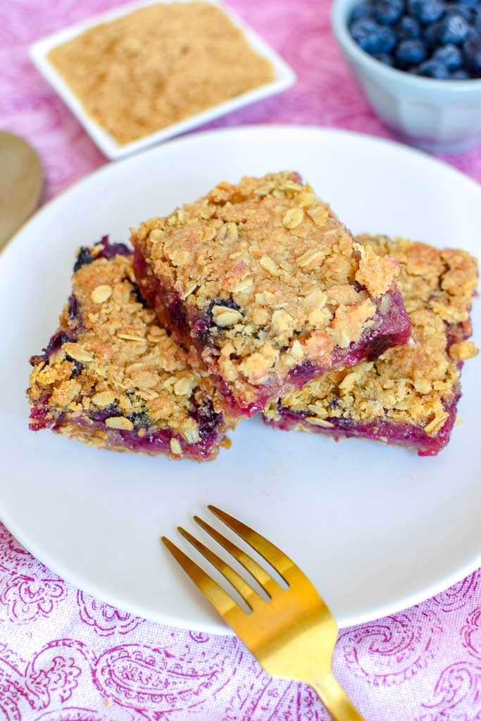 Berry, Rhubarb, and Oat Breakfast Bars shown on a white plate with a golden fork and a small dish of fresh blueberries and raw oats in the background.