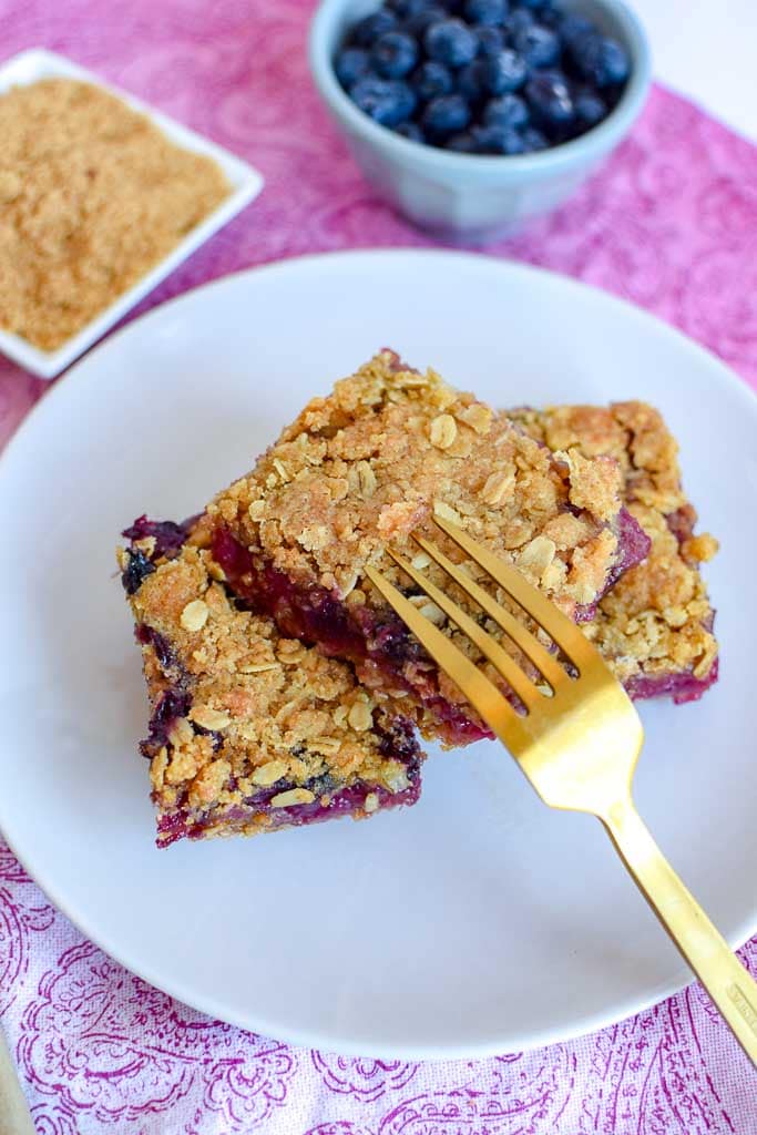 Berry, Rhubarb, and Oat Breakfast Bars shown on a white plate with a golden fork and a small dish of fresh blueberries in the background.