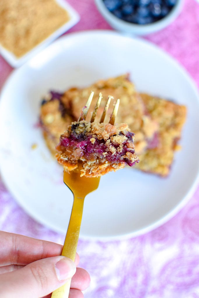 A bite of a breakfast bar made with strawberries, blueberries, and oats,  shown on a golden fork above the full breakfast bars on a plate.