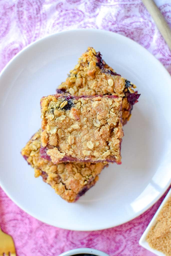 Three squares stacked of Berry, Rhubarb, and Oat Breakfast Bars shown on a white plate atop a pink tablecloth.