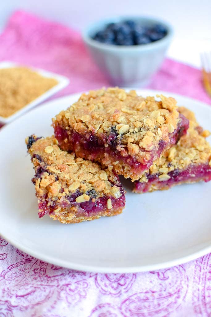 Berry, Rhubarb, and Oat Breakfast Bars shown on a white plate with a golden fork and a small dish of fresh blueberries in the background.