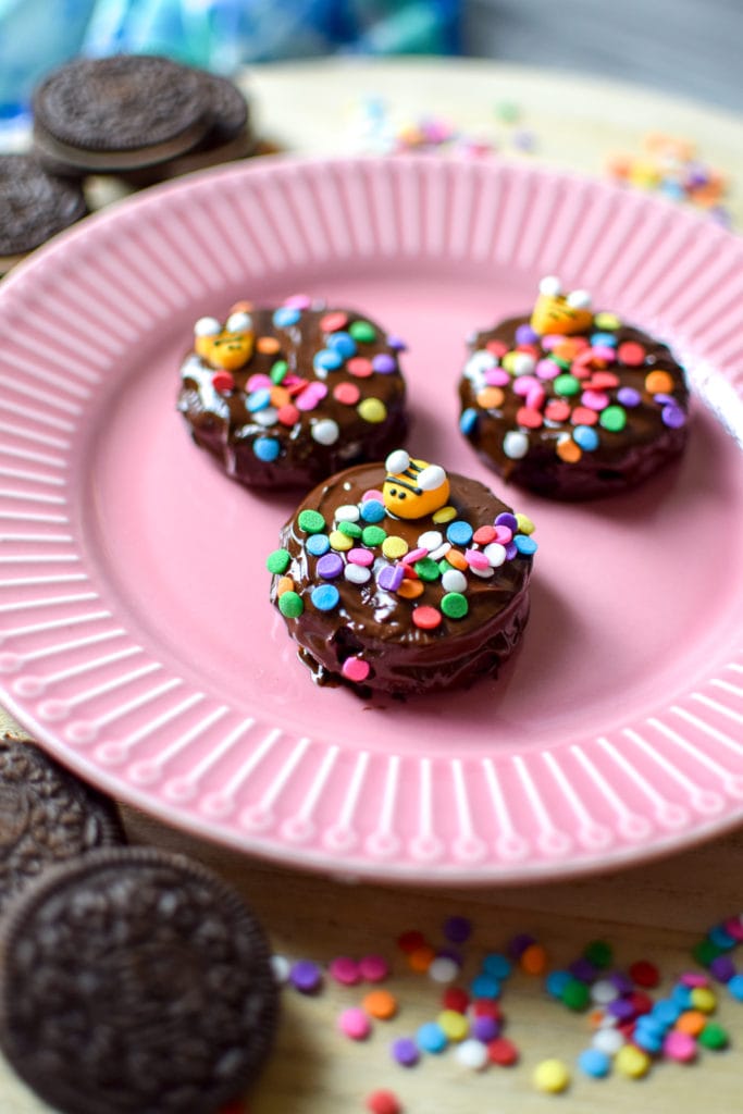 Three chocolate covered Oreos topped with sprinkles and a candy bee, on a pink plate surrounded by colorful sprinkles and Oreo cookies.