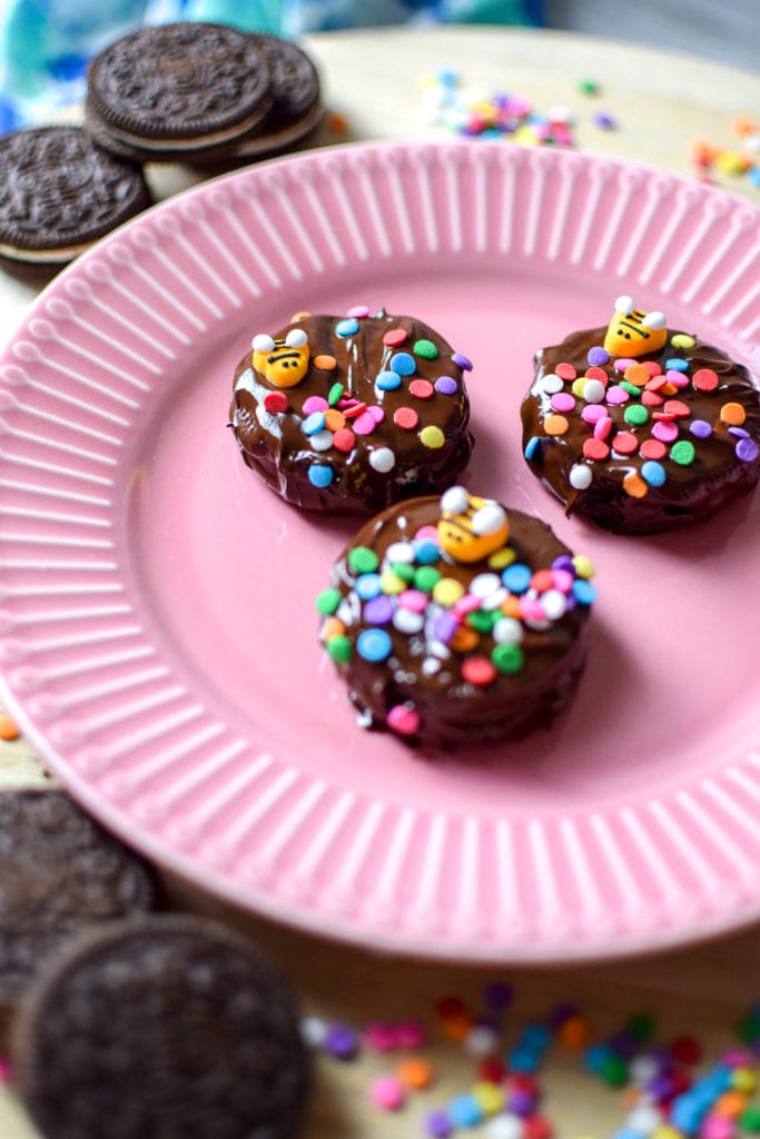 Chocolate covered Oreos on a pink plate surrounded by colorful sprinkles and Oreo cookies.