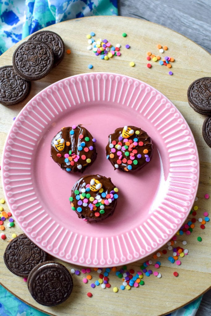 Three Finished Chocolate covered Oreos on a pink plate and wooden serving platter shown  with sprinkles and candy bees on top.