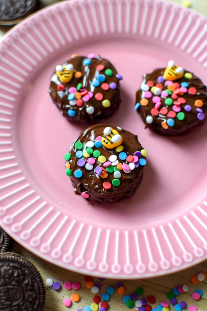 Chocolate covered oreos on a pink plate surrounded by colorful sprinkles and oreo cookies.