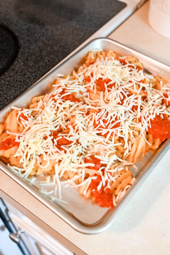 Waffle fries getting topped with shredded mozzarella cheese.