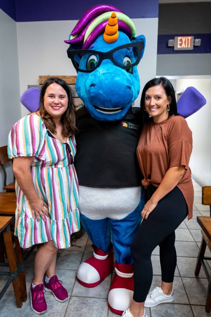 Sisters posing with the Delaware Public Library unicorn mascot in Fresh Start Bakery. 