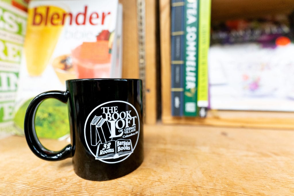 Black coffee mug with The Book Loft logo, sitting on a shelf at The Book Loft in German Village.