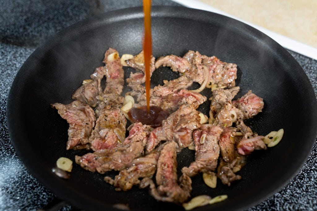 Flank steak being cooked in a pan on the stove with the marinade being poured over the meat. 