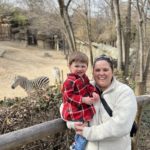 Mother in a white jacket holding her son in red plaid in front of a Zebra exhibit.