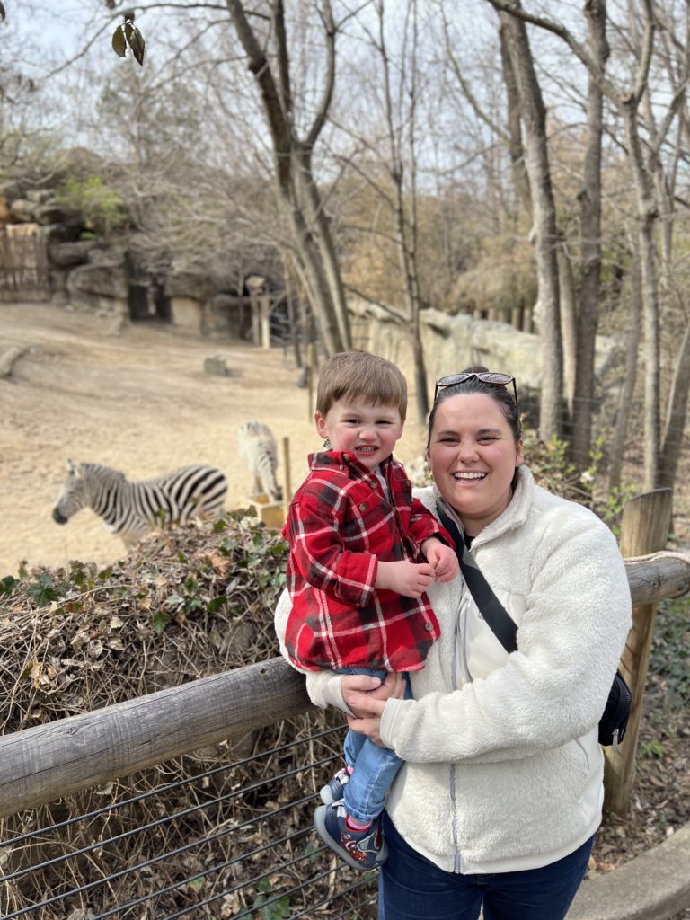 Mother in a white jacket holding her son in red plaid in front of a Zebra exhibit.