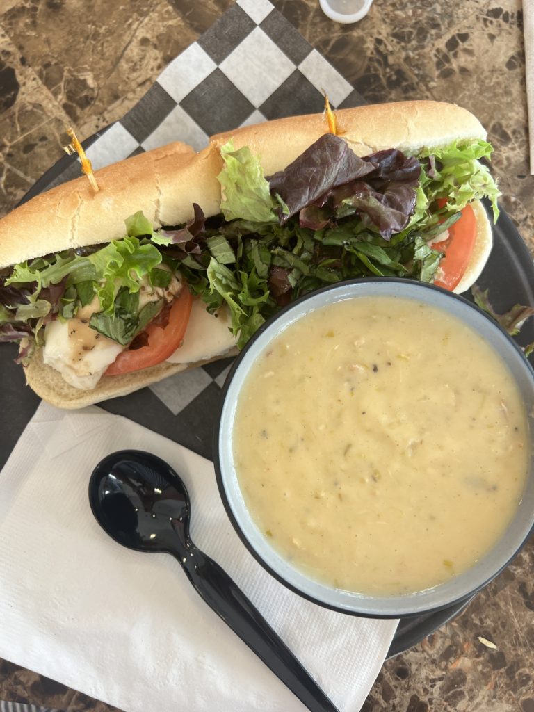 a bowl of she crab soup with a caprese sub on a wooden table with a white napkin and black plastic spoon.
